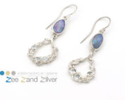 Zilveren (925) oorbellen bestaande uit getwijnde draden waar tussen natuurstenen topaas (licht blauw) gezet zijn. Onder het oor hangt een opaal gezet in zilveren zetkastje.